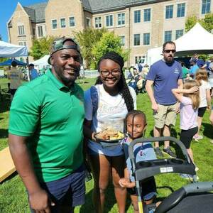 A man in a green polo shirt, a teenage girl holding a plate of food, and a small child in a stroller smile on the lawn in front of Dunne Hall.
