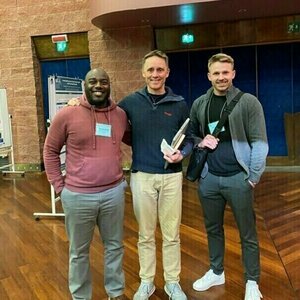 Three men smile for a photo in a hallway with wood floors and brick and blue walls.  The man on the left wears a maroon sweatshirt and gray pants.  The center man wears a dark gray quarter-zip and khaki pants.  The man on the right wears a gray zip-up jacket and dark gray pants.