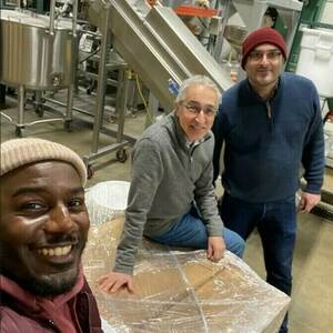 Three men, two in light sweaters and jeans, one in a maroon jacket and knit cap, pose around a large shrink-wrapped box in a warehouse with stainless steel equipment.