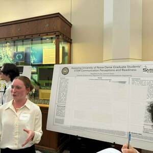 A woman wearing a white button-down shirt and a name tag gestures with her hands as she discusses her research poster titled "Assessing University of Notre Dame Graduate Students' STEM Communication Perceptions and Readiness."  Another student stands beside her by their poster, and a display case with scientific images is visible in the background.